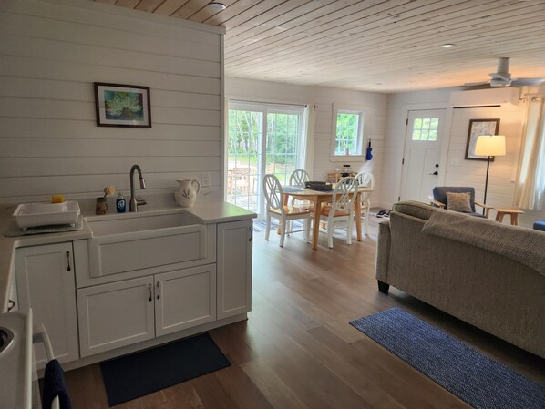 Una cocina moderna con armarios blancos, un comedor con mesa y sillas de madera, y una sala de estar con sofá y lámpara.