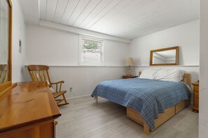 A bedroom with a wooden bed, a rocking chair, a bedside table, a lamp, and a window with blinds.