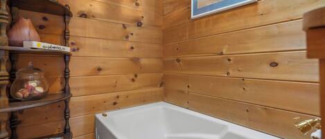 A wooden-paneled bathroom with a white bathtub, shelves with decorative items, and a framed picture on the wall.