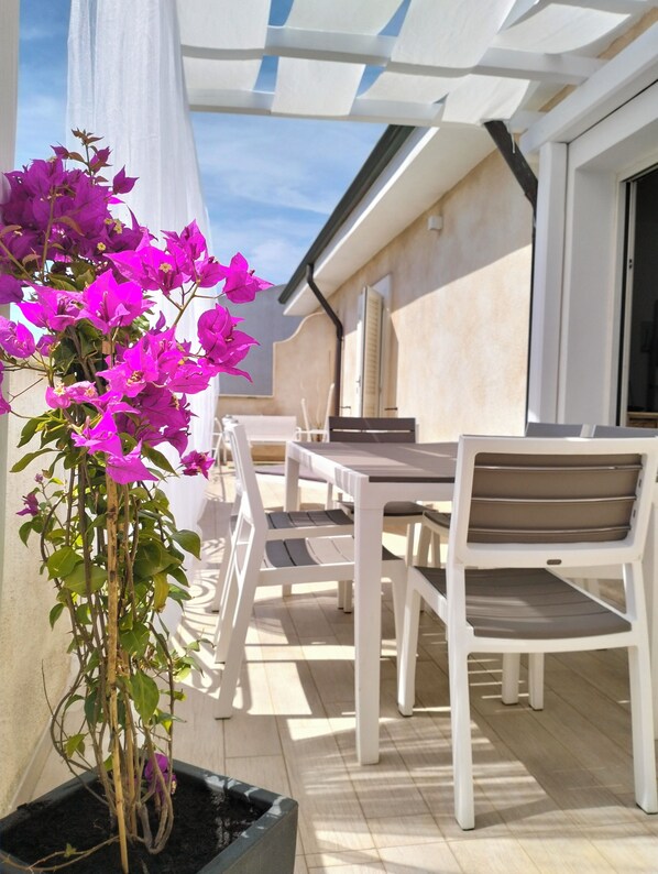 An outdoor patio with a table and chairs, a potted plant with purple flowers, and a white awning providing shade.