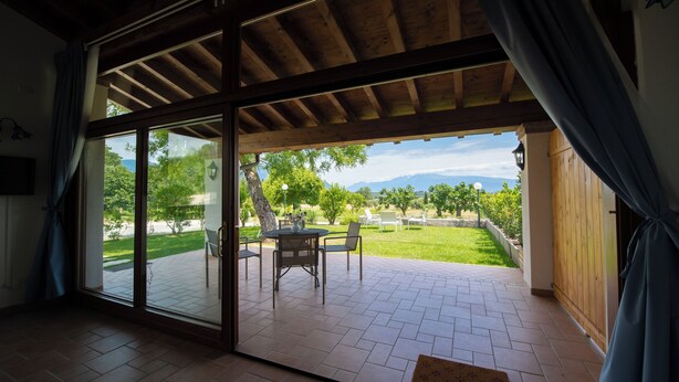 A spacious room with a large sliding glass door leading to an outdoor patio area with a table and chairs, a view of greenery and mountains, and a wooden ceiling. - Il Podere del Bardo (Puegnago del Garda)