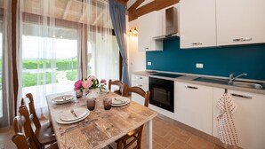 A dining area with a wooden table set for four, a kitchen with white cabinets and a blue backsplash, and a view of greenery through the window. - Il Podere del Bardo (Puegnago del Garda)
