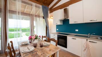 Uma área de refeições com mesa de madeira posta para quatro, uma cozinha com armários brancos e painel azul, e vista para o verde pela janela.