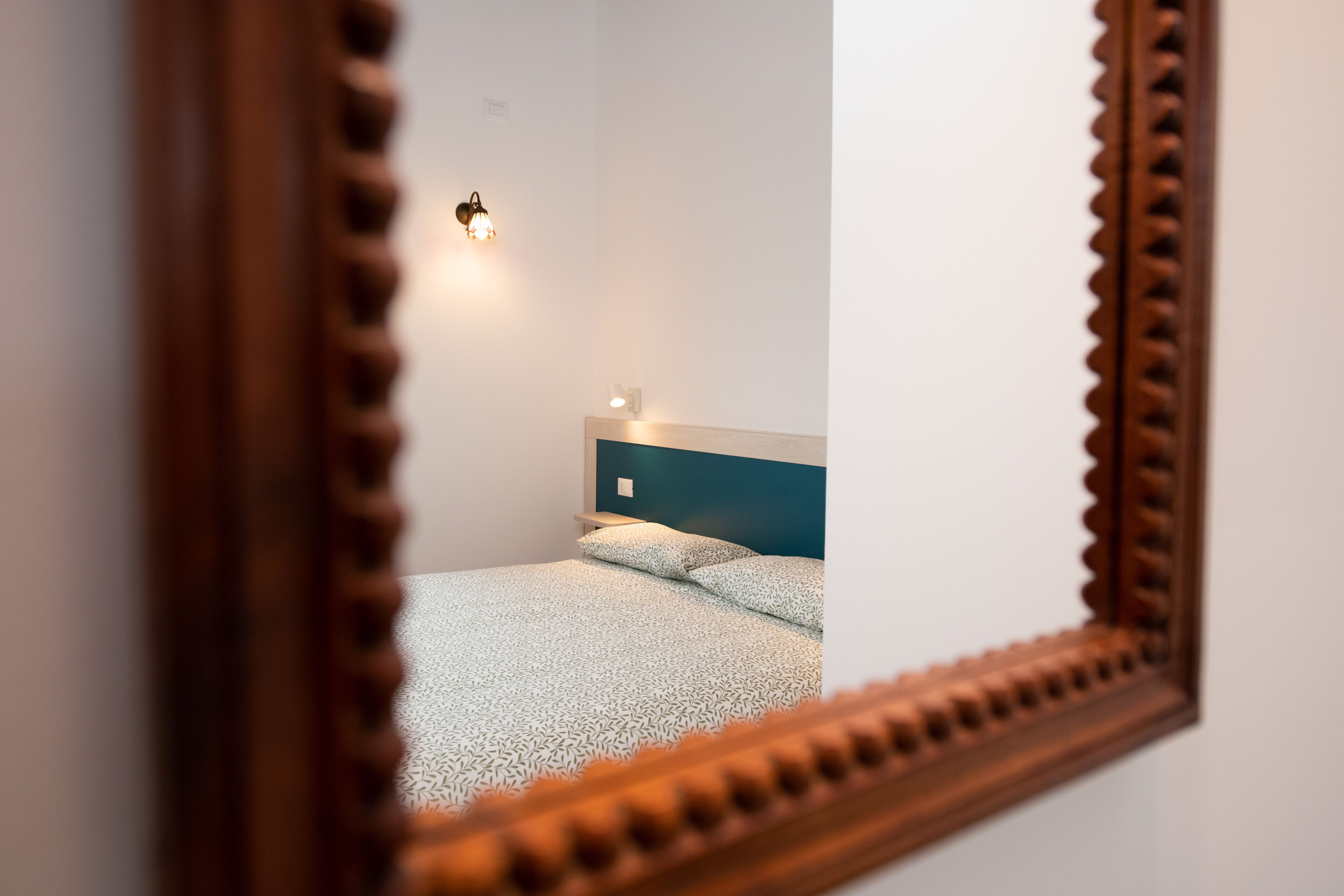 A bedroom with a bed, a nightstand, a wall-mounted light, and a mirror with a wooden frame.