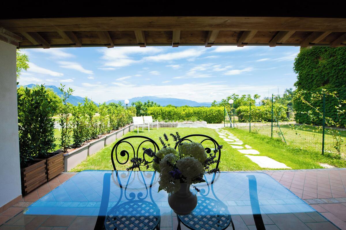 A patio with a table and chairs, a vase with flowers, and a view of a garden and mountains.