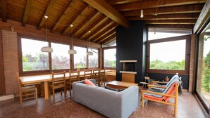 Amplia sala de estar con techo de madera, ventanas grandes, un sofá gris, sillas de madera, una mesa de comedor y un televisor.