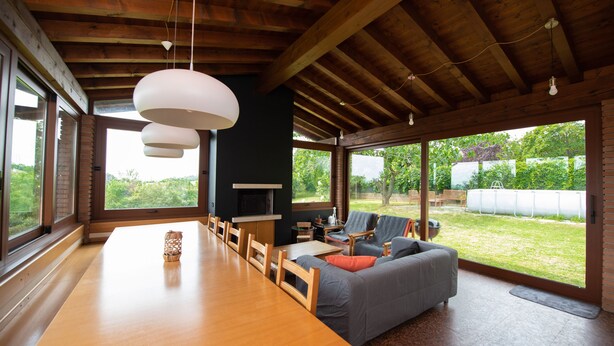 Una habitación amplia con una larga mesa de madera, un sofá gris con cojines naranja y una chimenea. Ventanas grandes ofrecen vistas a un jardín.