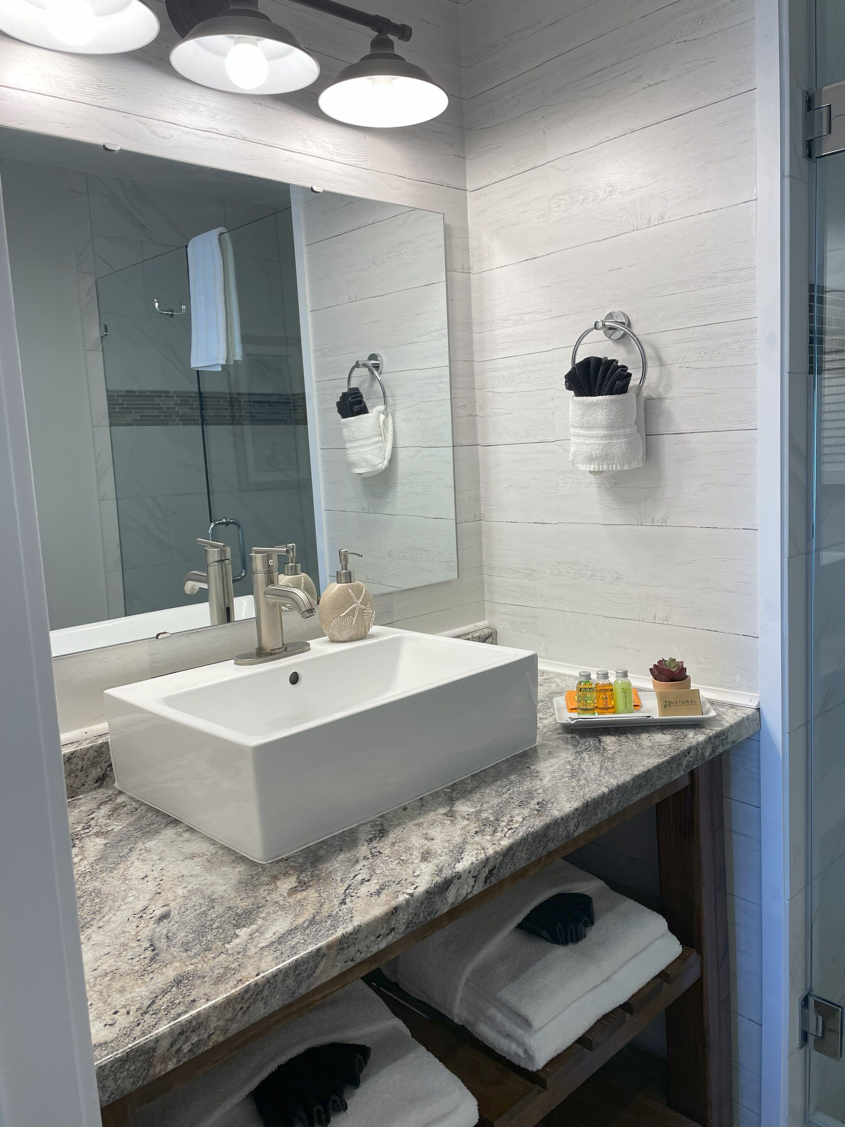 A modern bathroom with a large mirror, a white sink, and a granite countertop. There are two towel racks with white towels and a small plant on the countertop.