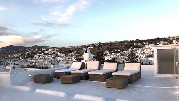 Une terrasse sur le toit, dotée de chaises longues et d’un espace salon couvert, offrant une vue sur une ville côtière aux bâtiments blancs et aux collines en arrière-plan.