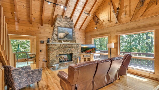 A wooden cabin interior with a stone fireplace, leather sofa, and large windows. -  Log cabin between Boone and Banner Elk-hot tub-game room-fire pit-private-cozy (Banner Elk)