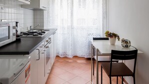A compact kitchen with a microwave, stove, and cabinets, a dining table with chairs, and a window with a white curtain. - PennyLane Apartment 1 (Roma)