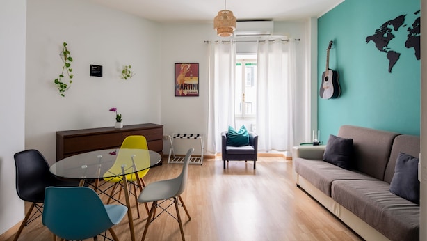 A modern living room with a sofa, a dining table with chairs, a small kitchen area, and a view of the outdoors. - PennyLane Apartment 1 (Roma)