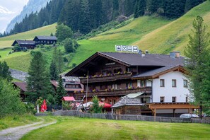 Un bâtiment traditionnel en bois de style alpin, avec une enseigne indiquant qu’il s’agit d’un restaurant, entouré d’une végétation luxuriante et de montagnes.