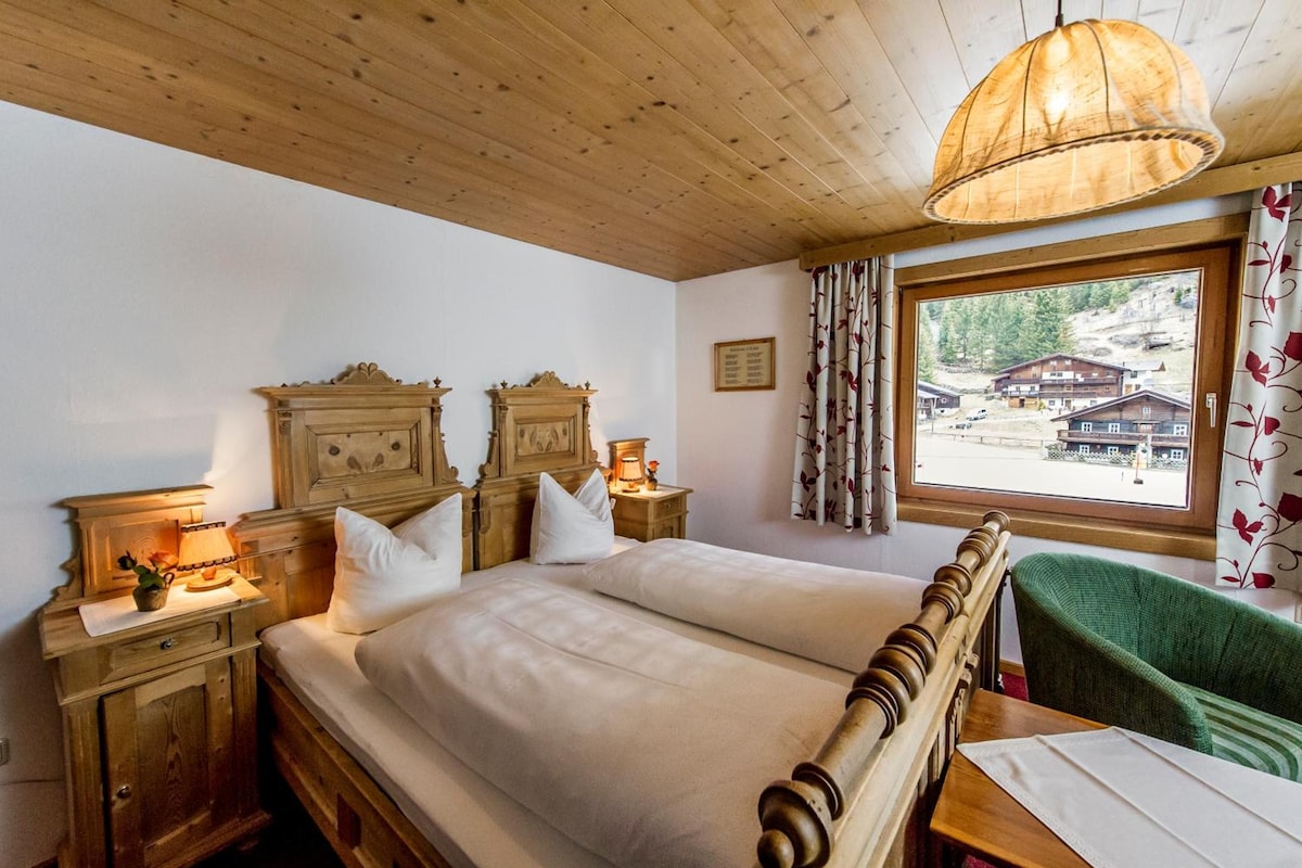 A room with a wooden ceiling, a large bed with white linens, two bedside tables with lamps, a window with a view of buildings, and a green chair.