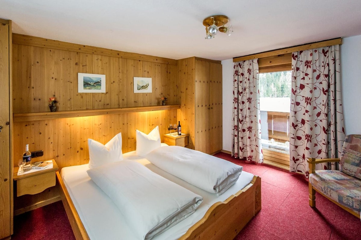 A wooden-paneled bedroom with a double bed, bedside tables, a chair, and a window with curtains.
