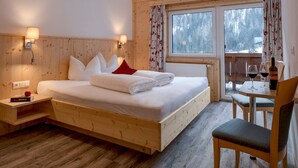 A wooden bedroom with a bed, bedside tables, a window with curtains, and a dining area with a chair and table. - Zollwirt (Sankt Jakob in Defereggen)
