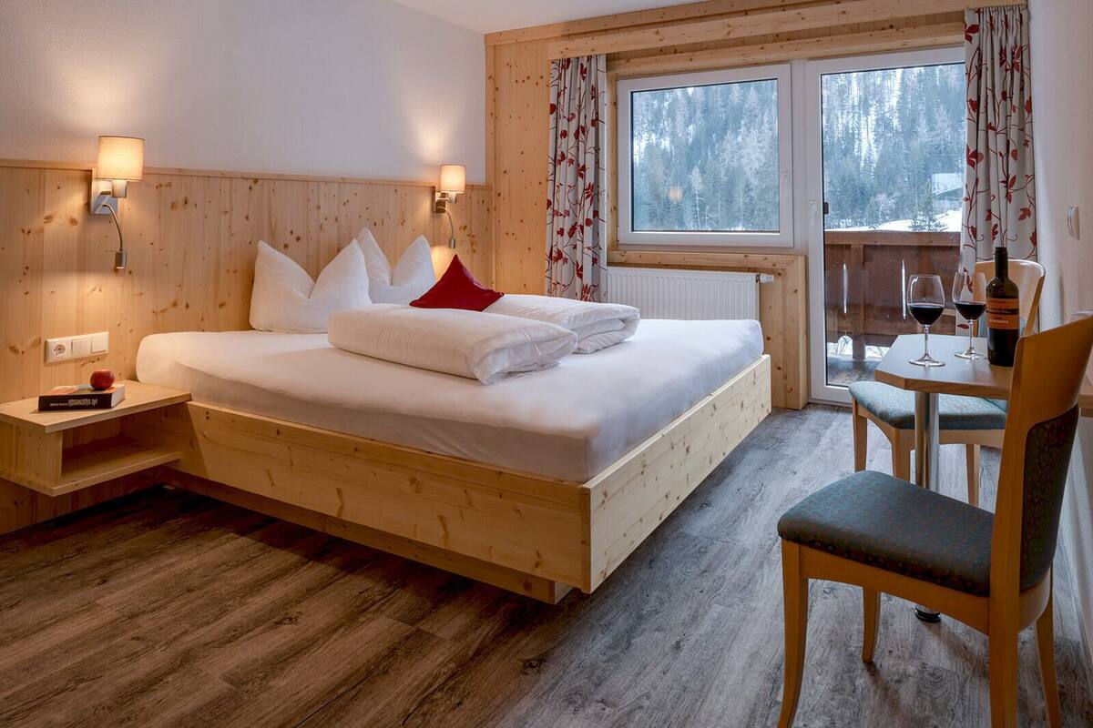 A wooden bedroom with a bed, bedside tables, a window with curtains, and a dining area with a chair and table.