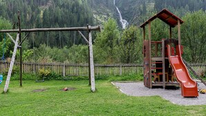A playground with a slide and swings, surrounded by a forested area and a waterfall in the background. - Zollwirt (Sankt Jakob in Defereggen)