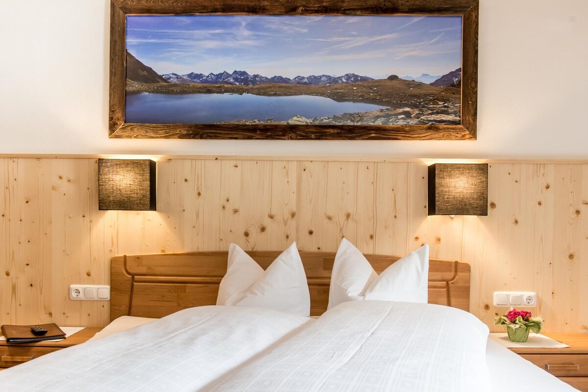 A hotel room with a wooden headboard, a large bed with white bedding, two bedside tables with lamps, and a framed landscape painting above the bed.