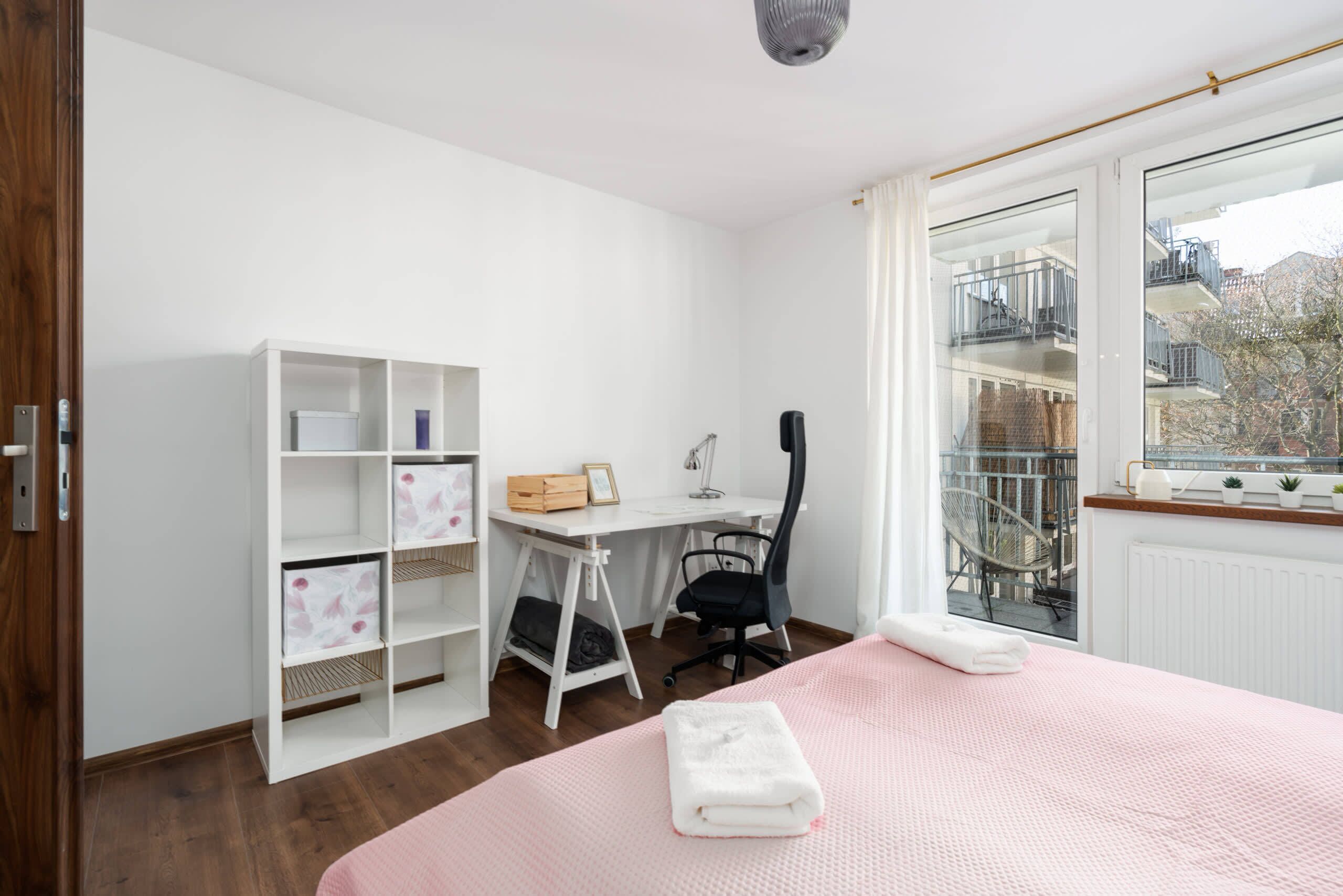 Une chambre à coucher comprenant un lit, un bureau, une chaise et une fenêtre donnant sur des immeubles.