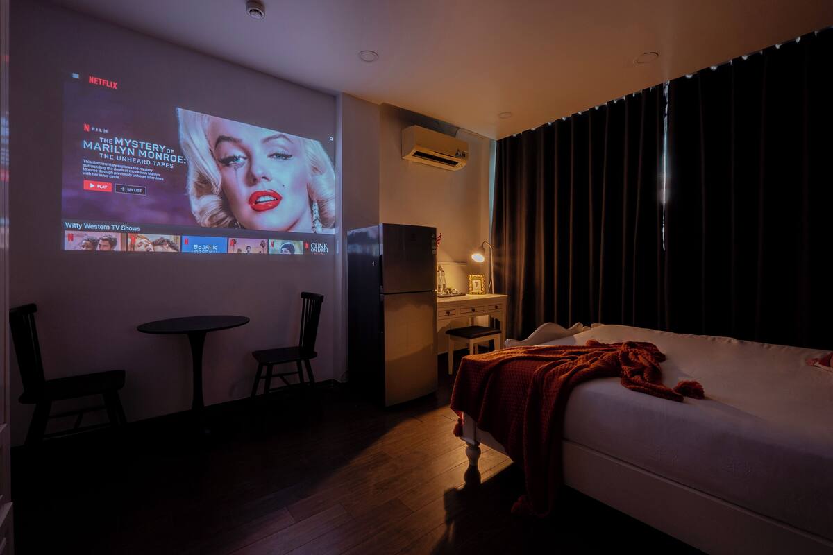 A hotel room with a bed, a small table, a chair, a TV mounted on the wall, and a window with curtains.