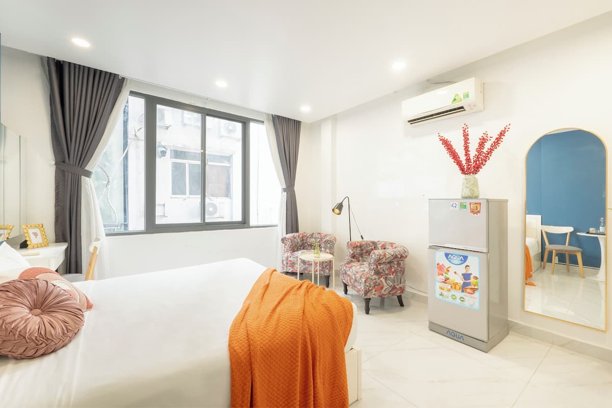A hotel room with a bed, two armchairs, a small table, an air conditioning unit, and a refrigerator with a water dispenser.