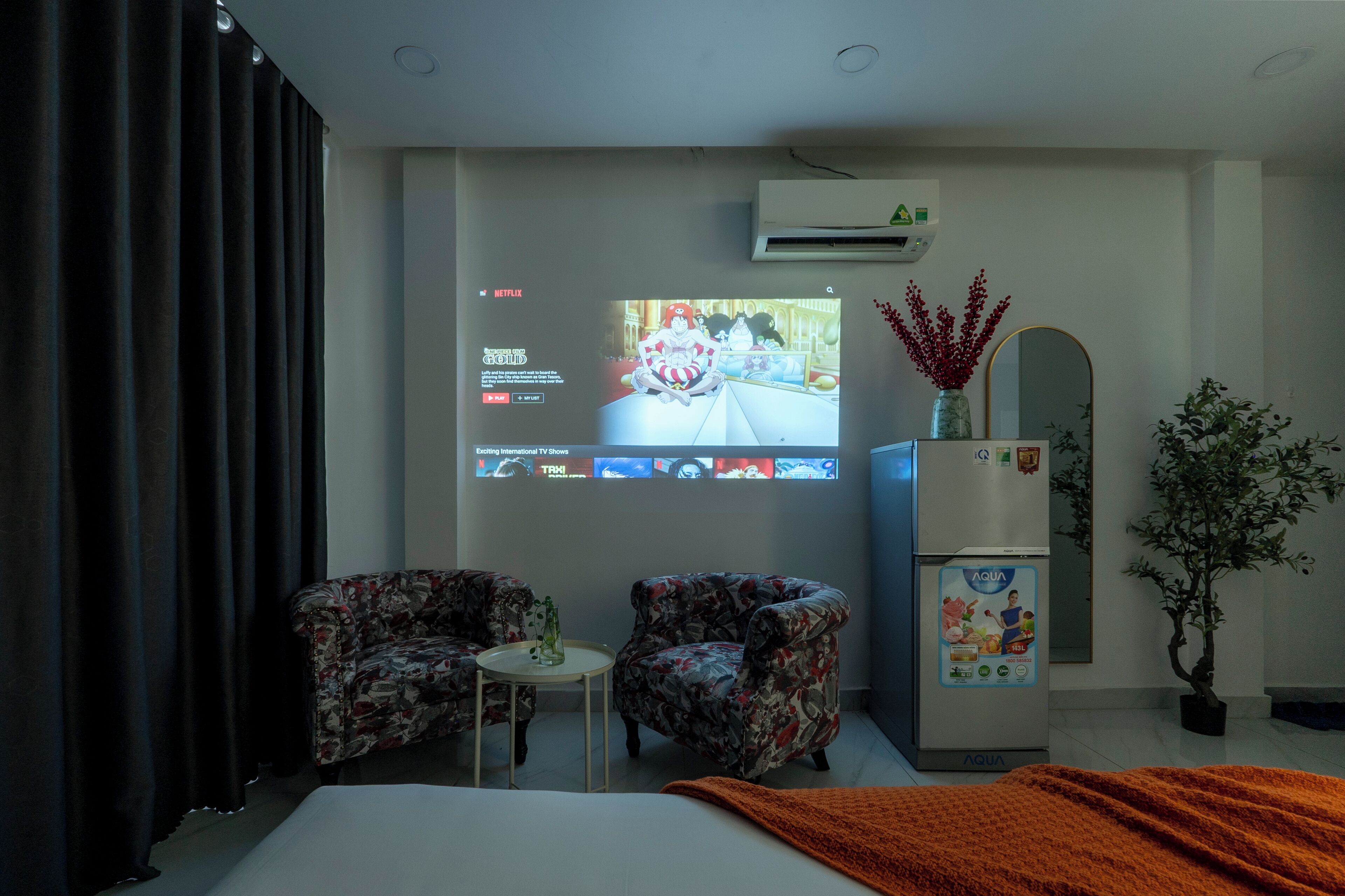 Ein Hotelzimmer mit einem Bett, zwei blumigen Sesseln, einem kleinen Tisch, einem Fernseher, der eine Sendung zeigt, einer Klimaanlage, einem Kühlschrank und einer Topfpflanze.