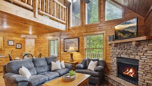 A cozy cabin interior with a stone fireplace, wooden walls, and a large window overlooking greenery. - Relax Above the Smokies · Bearway to Heaven (Sevierville)