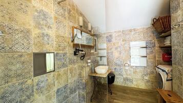 Un baño con baldosas estampadas, un lavamanos, un espejo, un perchero para toallas, una repisa con una cesta y una lavadora.