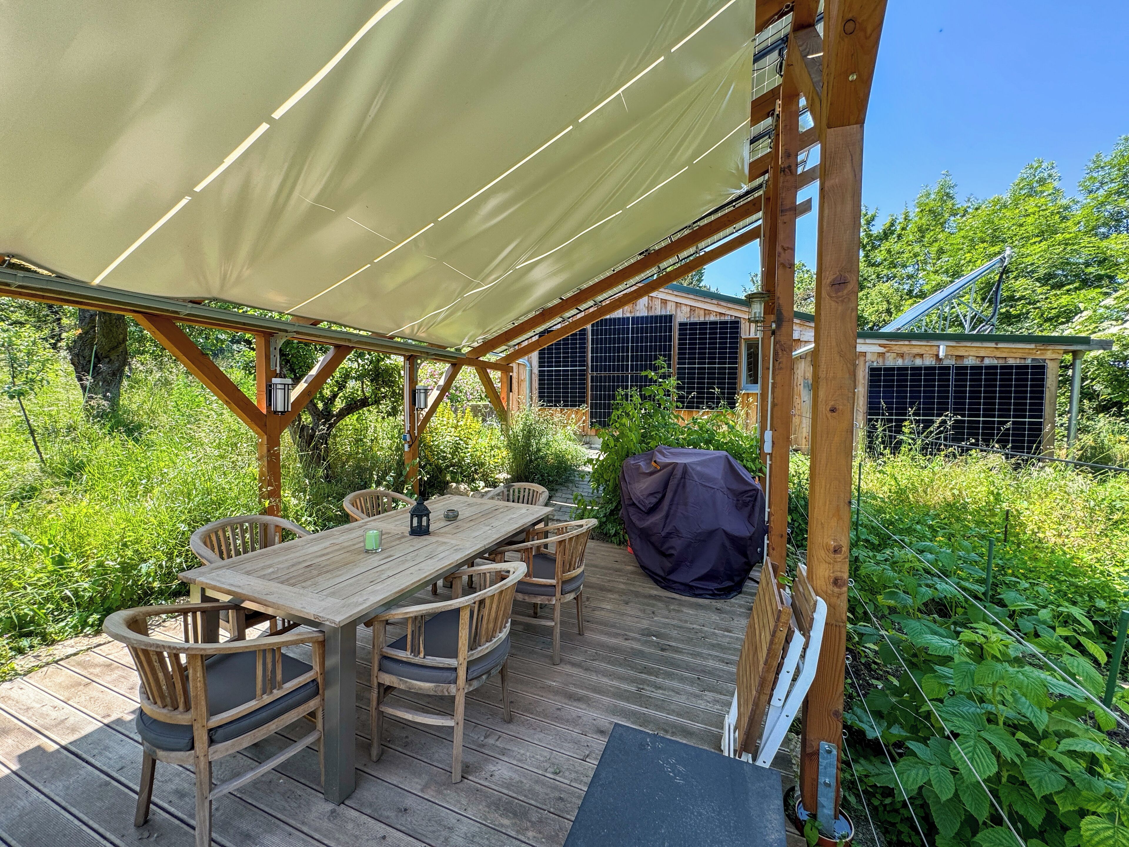 Une terrasse en bois avec un espace patio couvert, des sièges d’extérieur et, au fond, une structure abritant des panneaux solaires.