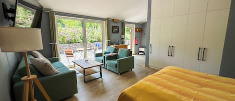 A modern bedroom with a large bed, a sofa, a coffee table, and a view of the garden through the sliding doors.