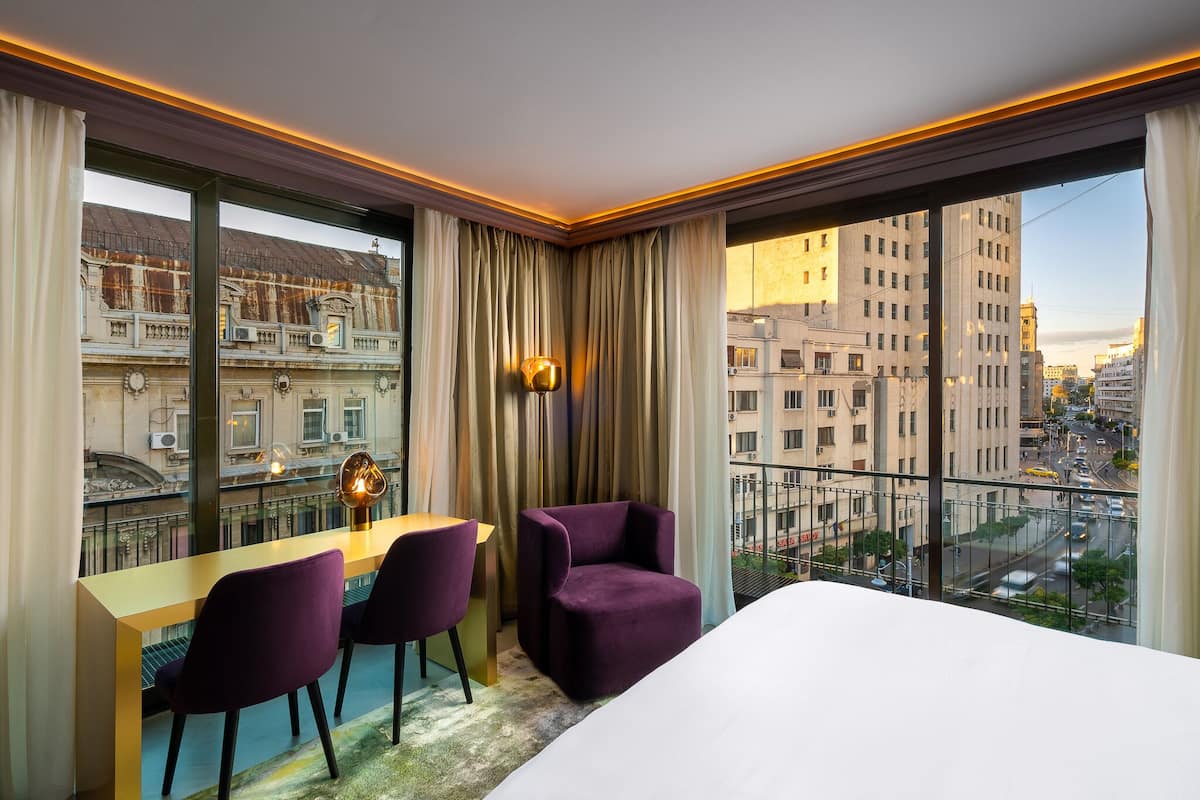 A hotel room with a large window, a desk with two chairs, a bed, and a view of the cityscape.