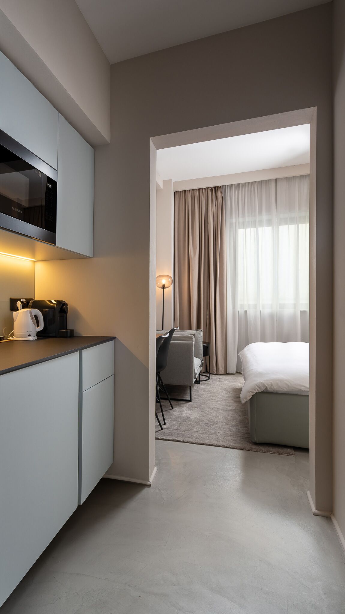 Una habitación de hotel moderna con kitchenette, cama, escritorio y un ventanal con cortinas.