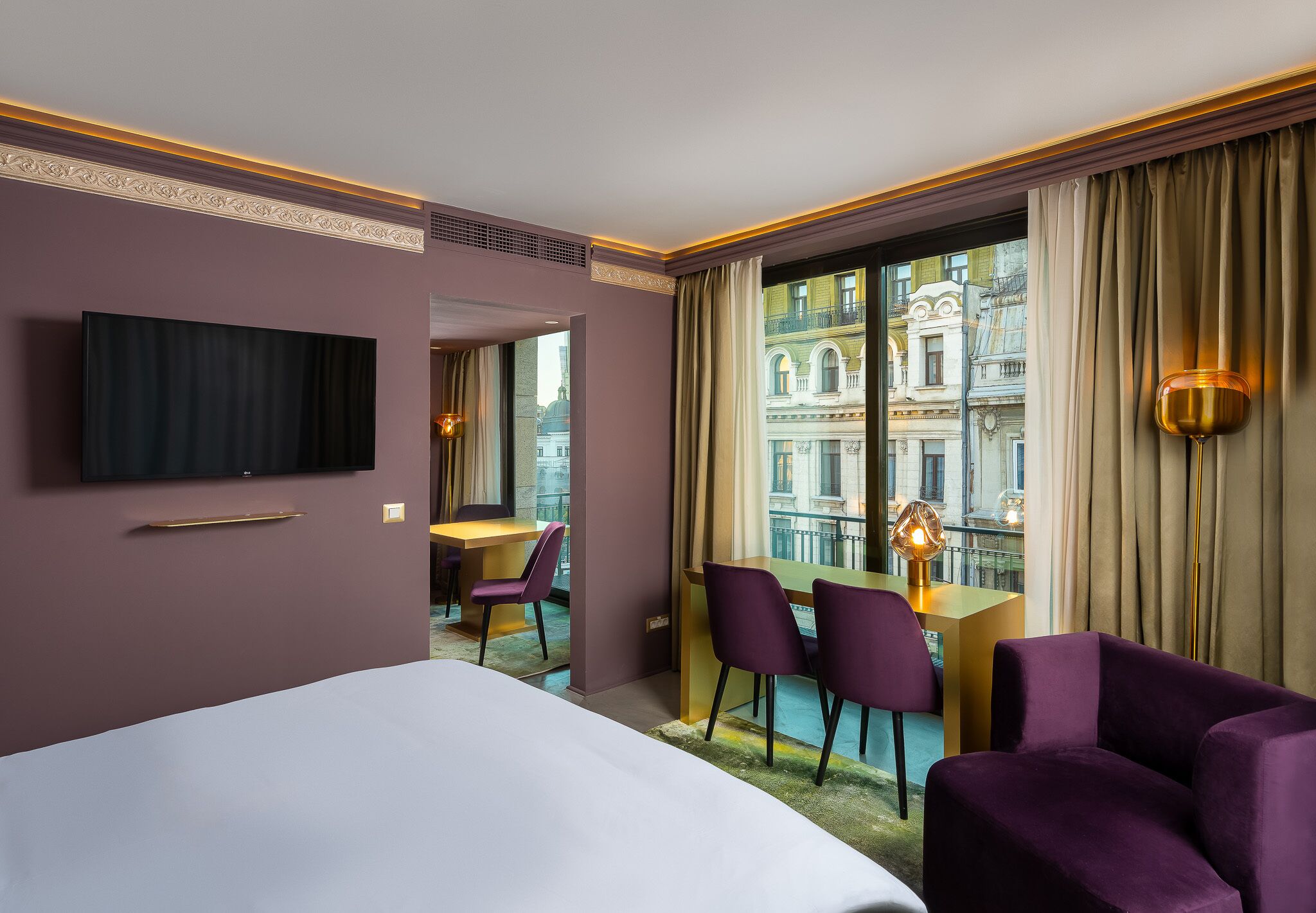 Une chambre d’hôtel avec un lit, un bureau, des chaises violettes, une télévision et une vue sur les immeubles à travers de grandes fenêtres.