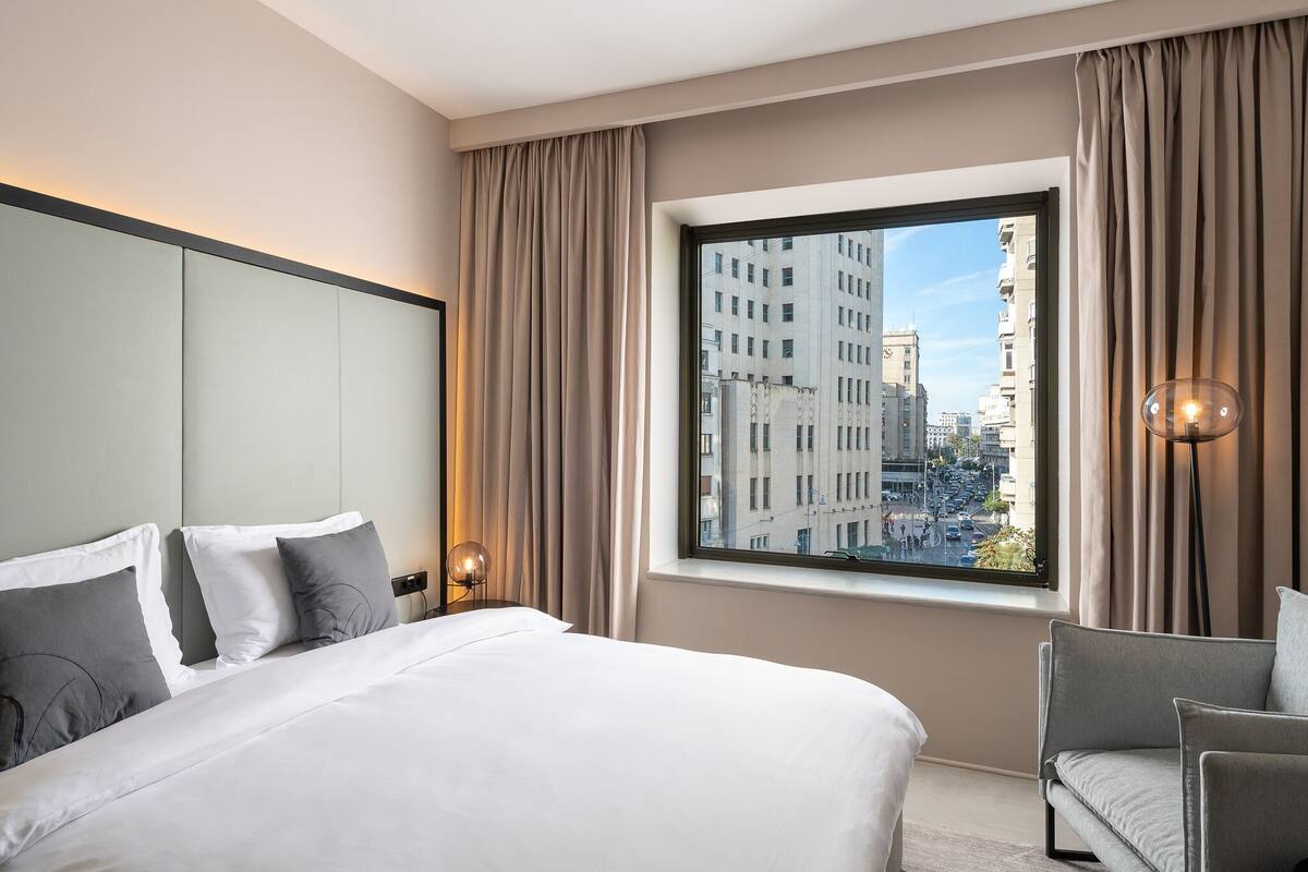 A hotel room with a large bed, a view of the city through a window, and a chair.