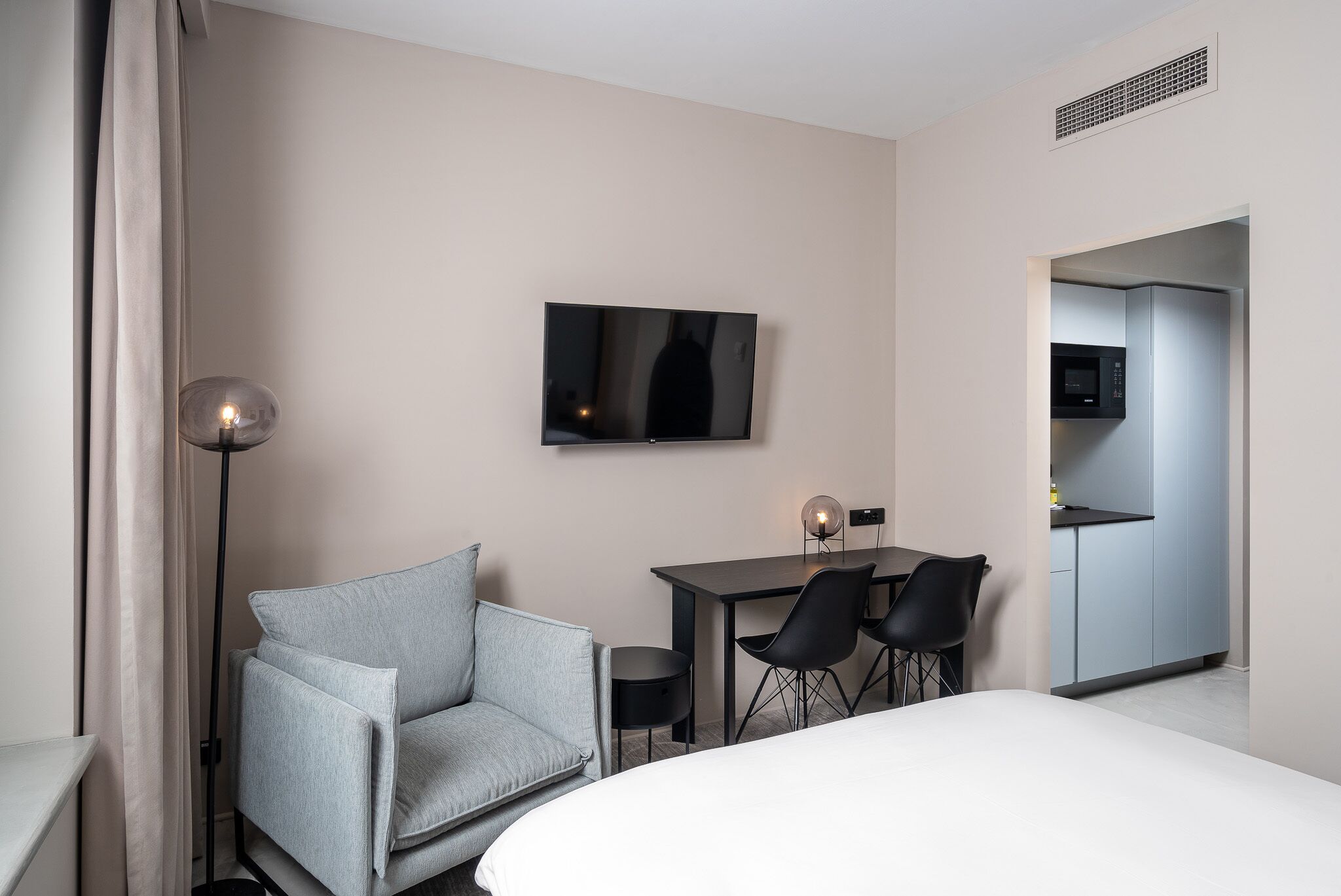 A modern hotel room with a bed, a grey armchair, a desk with two chairs, a flat-screen TV, and a kitchenette.