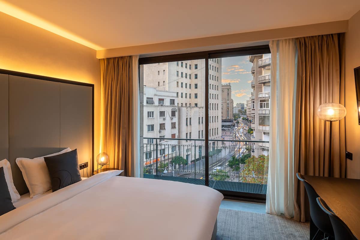 A hotel room with a large bed, a desk, and a view of the city through a balcony.