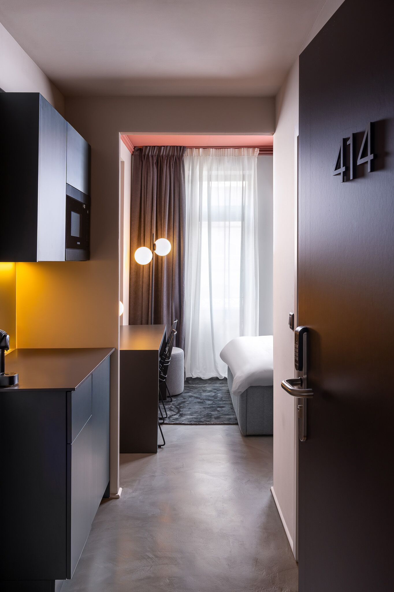 Quarto de hotel moderno com cozinha compacta, cama, escrivaninha e uma grande janela com cortinas.