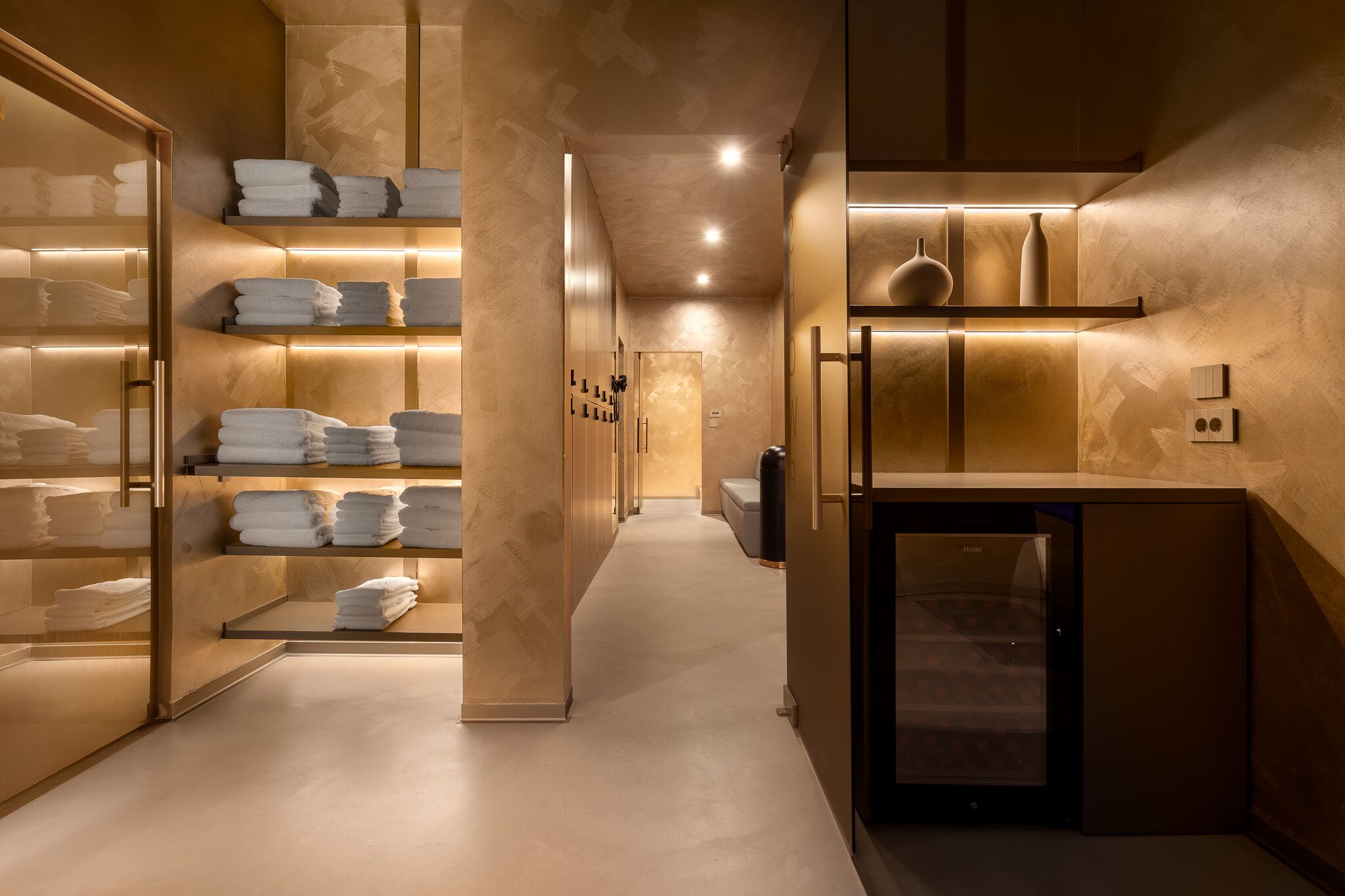 Una hall d'hotel moderna con un corridoio centrale, scaffali in legno con asciugamani piegati, una reception e una macchina per il caffè.
