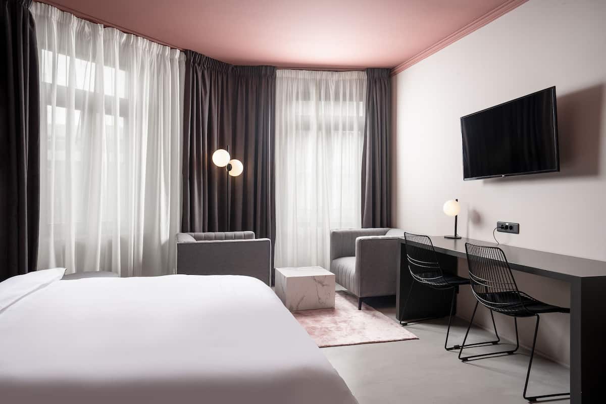 A modern hotel room with a large bed, two armchairs, a desk, and a TV.