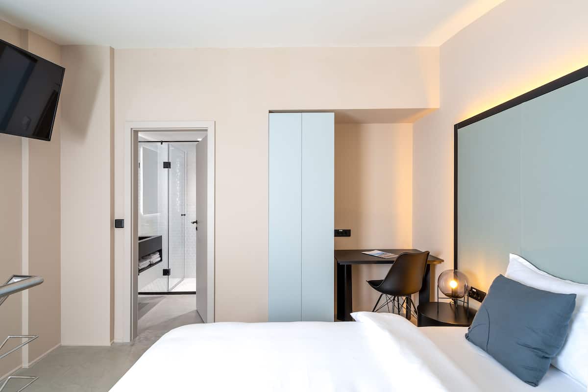 A modern hotel room with a bed, a desk, a chair, and a TV mounted on the wall.