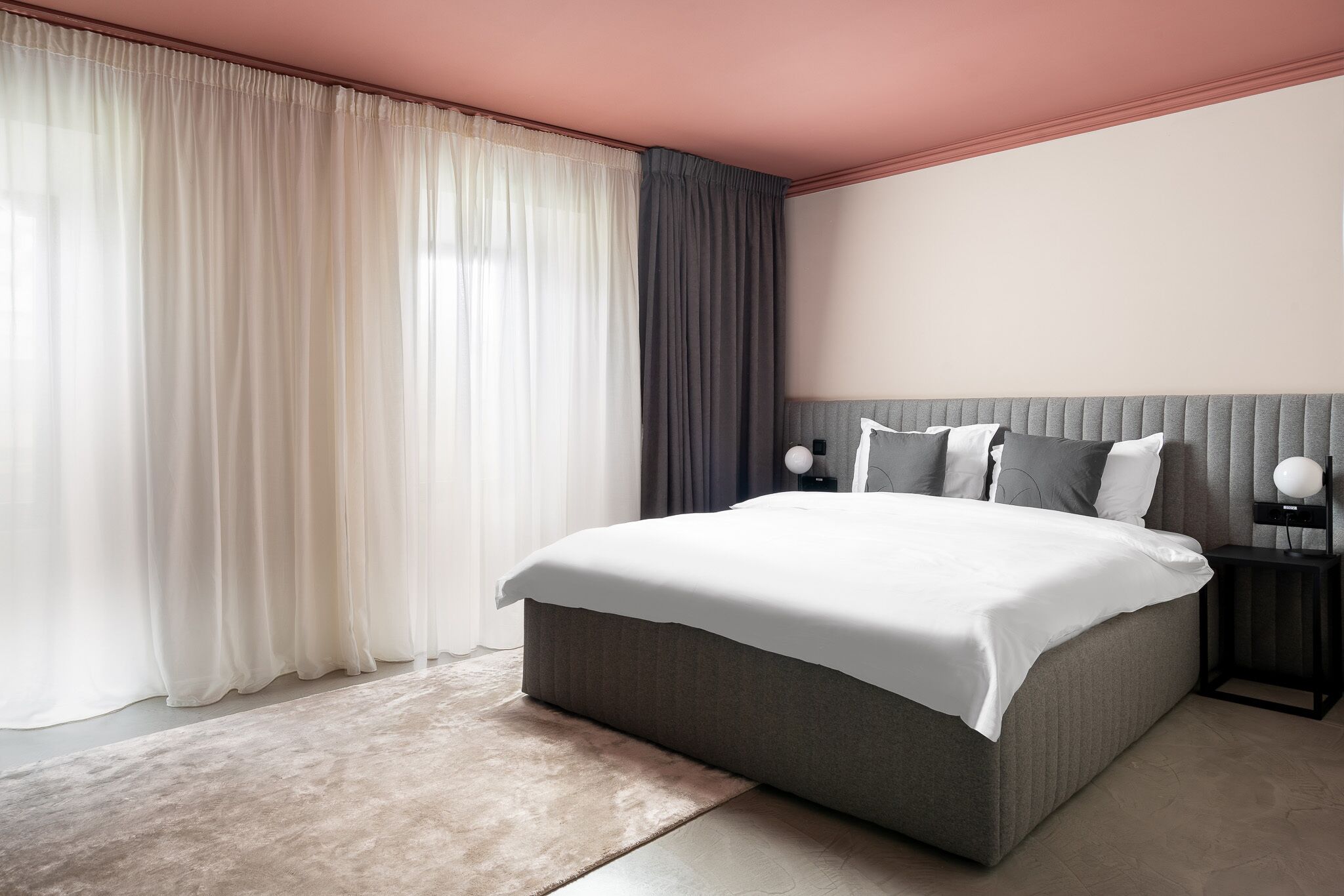 A modern bedroom with a large bed, a nightstand with a lamp, and sheer curtains on the window.
