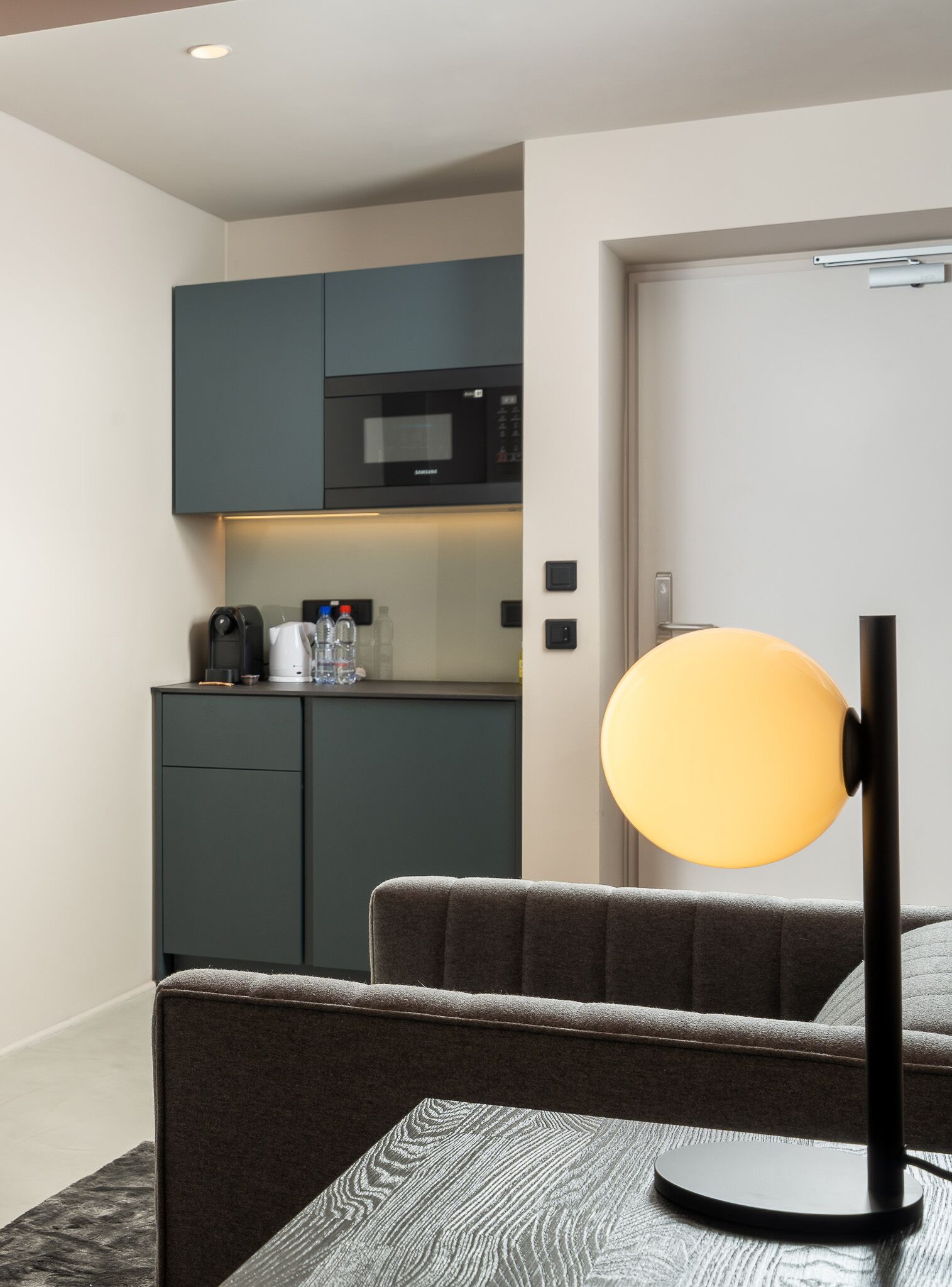 A compact hotel room with a kitchenette, a sofa, and a floor lamp.