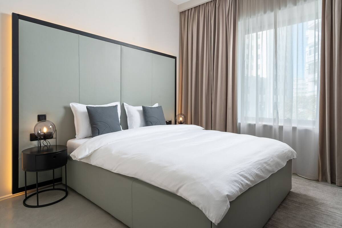 A modern bedroom with a large bed, a nightstand with a lamp, and a view of the city through sheer curtains.
