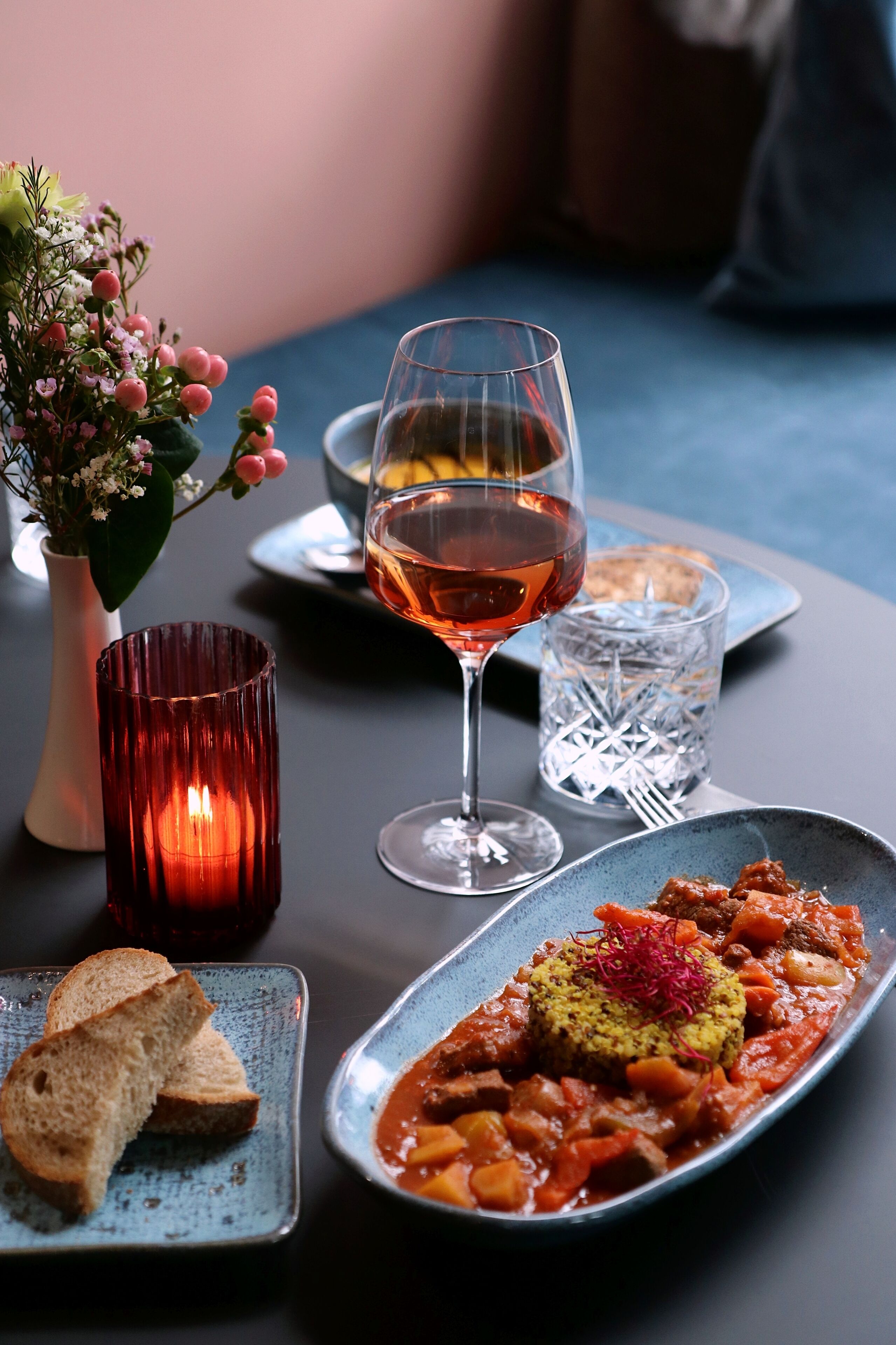 A dining table with a plate of food, a glass of red wine, a candle, and a vase of flowers.