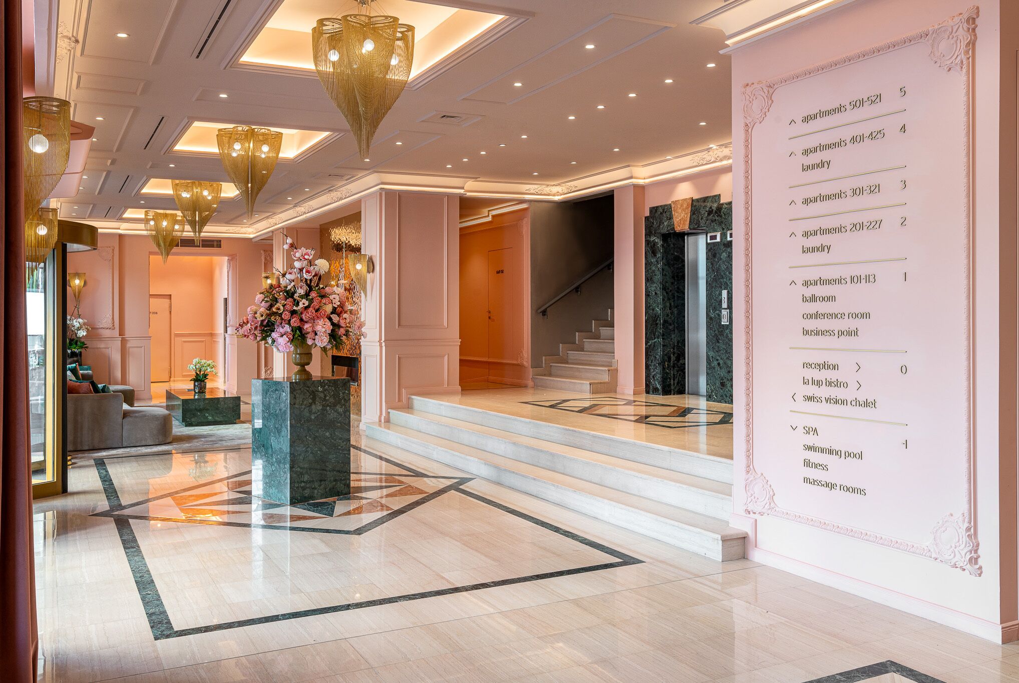A hotel lobby with a marble floor, a reception desk, a staircase, and a wall with a directory listing amenities like apartments, laundry, and a spa.