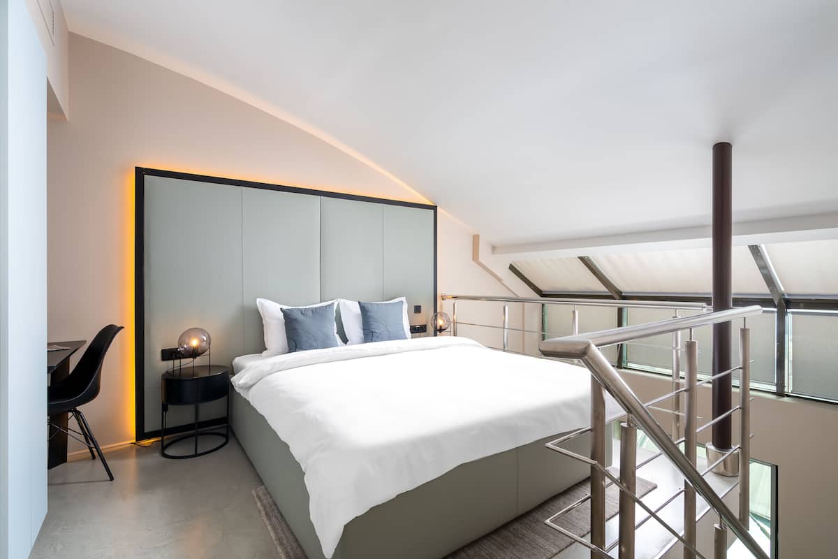 A modern bedroom with a large bed, a bedside table with a lamp, a chair, and a view of the sky through a balcony railing.