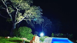 A nighttime view of a landscaped garden with a swimming pool, a large tree, and a well-lit pathway. - Villa Taglio Rosso (Sainte Lucie de Porto Vecchio)