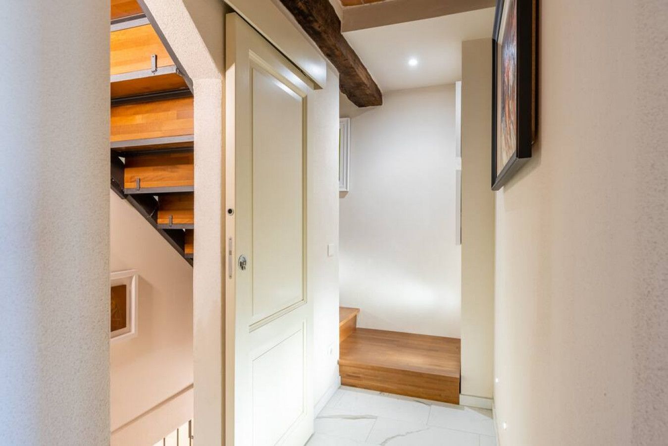 Un pasillo con una escalera de madera, una puerta blanca y un cuadro en la pared.