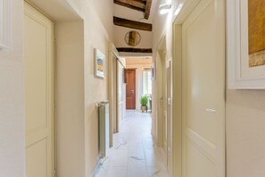 Ein Korridor mit cremefarbenen Wänden, einem Marmorboden und Holzbalken an der Decke. Auf beiden Seiten befinden sich Türen, und an der linken Wand ist ein Heizkörper zu sehen.
